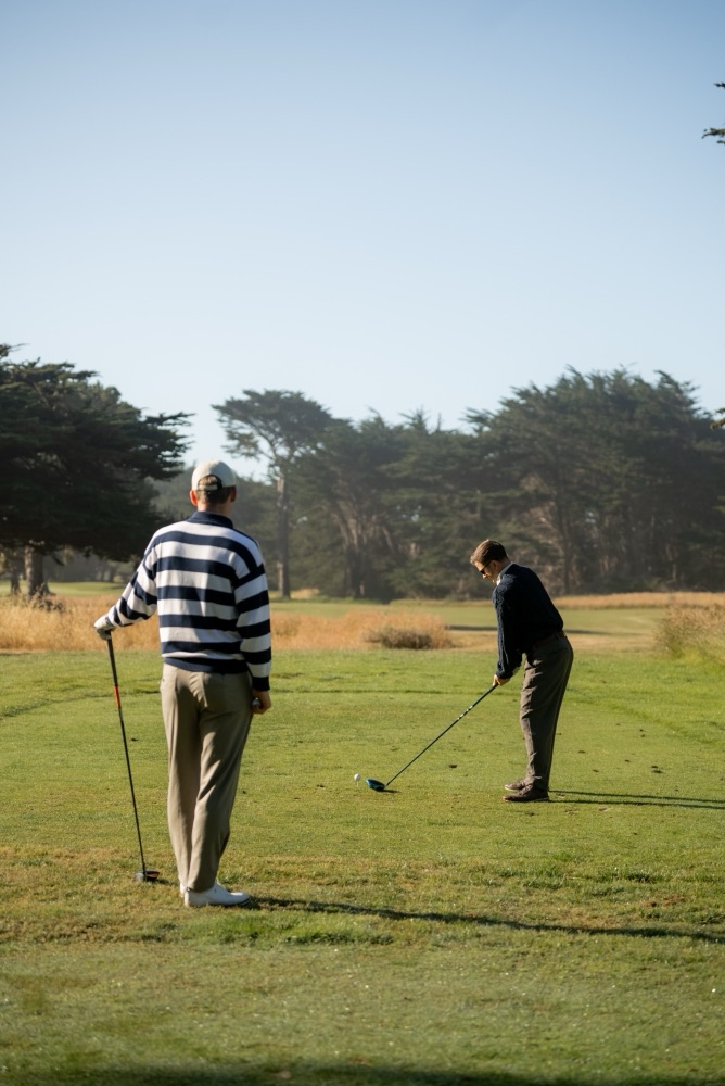 Golfer in a striped sweater watching a friend tee off among cypress trees