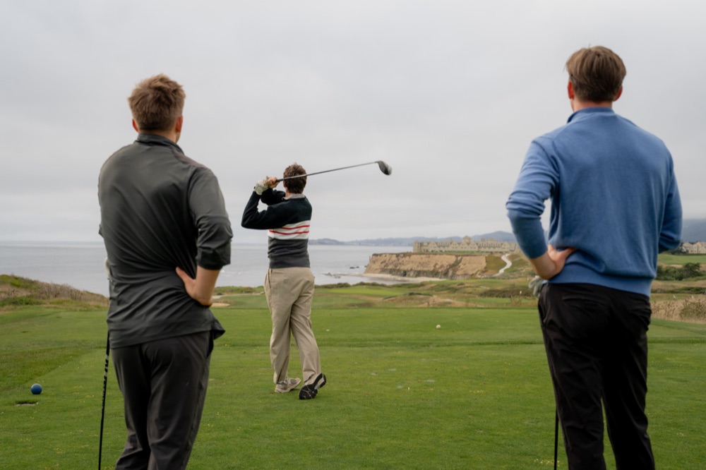 Two golfers watching a friend tee off on a clifftop course above the ocean